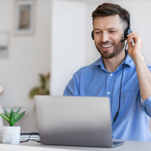 Man in dress shirt working at a desk with his hand on a headset, smiling, laptop in front of him.