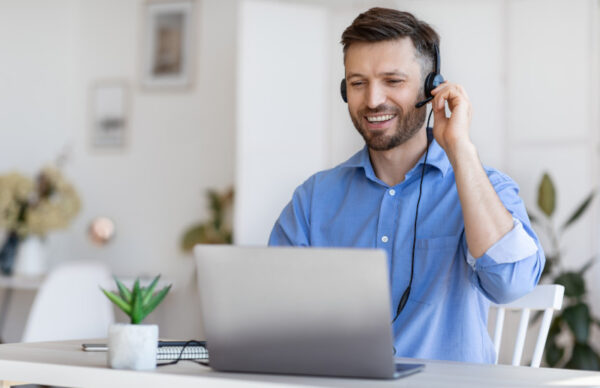 Man in dress shirt working at a desk with his hand on a headset, smiling, laptop in front of him.